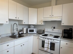A compact kitchen with white cabinets, a stainless steel sink, a microwave, a toaster, a stove, and a Keurig coffee maker. - Saltwater Steeple- Bell Tower Suite (Port Saunders)