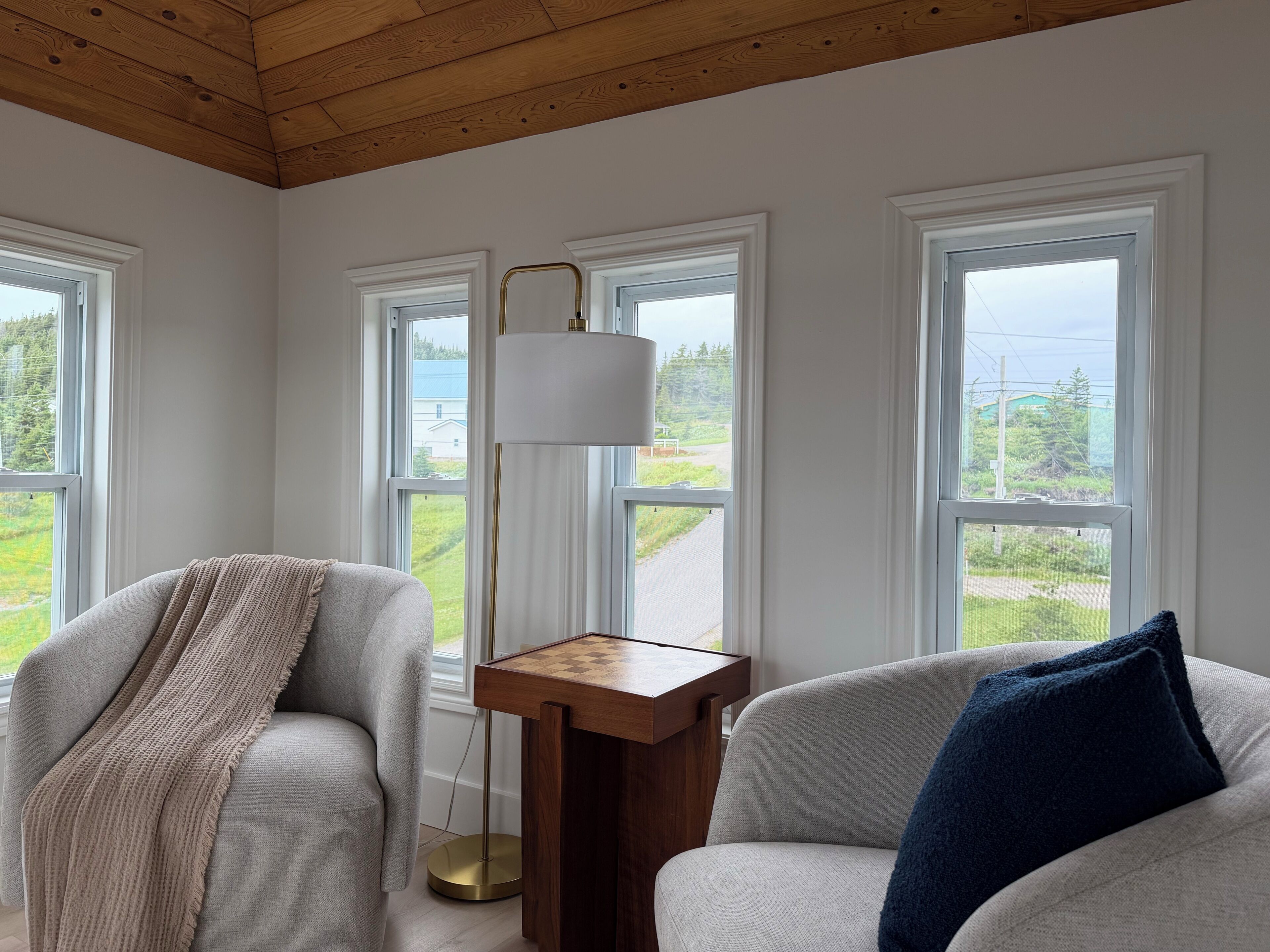 Une pièce avec un plafond en bois, deux fenêtres à cadres blancs, un canapé gris avec un plaid beige, une petite table en bois et un lampadaire.