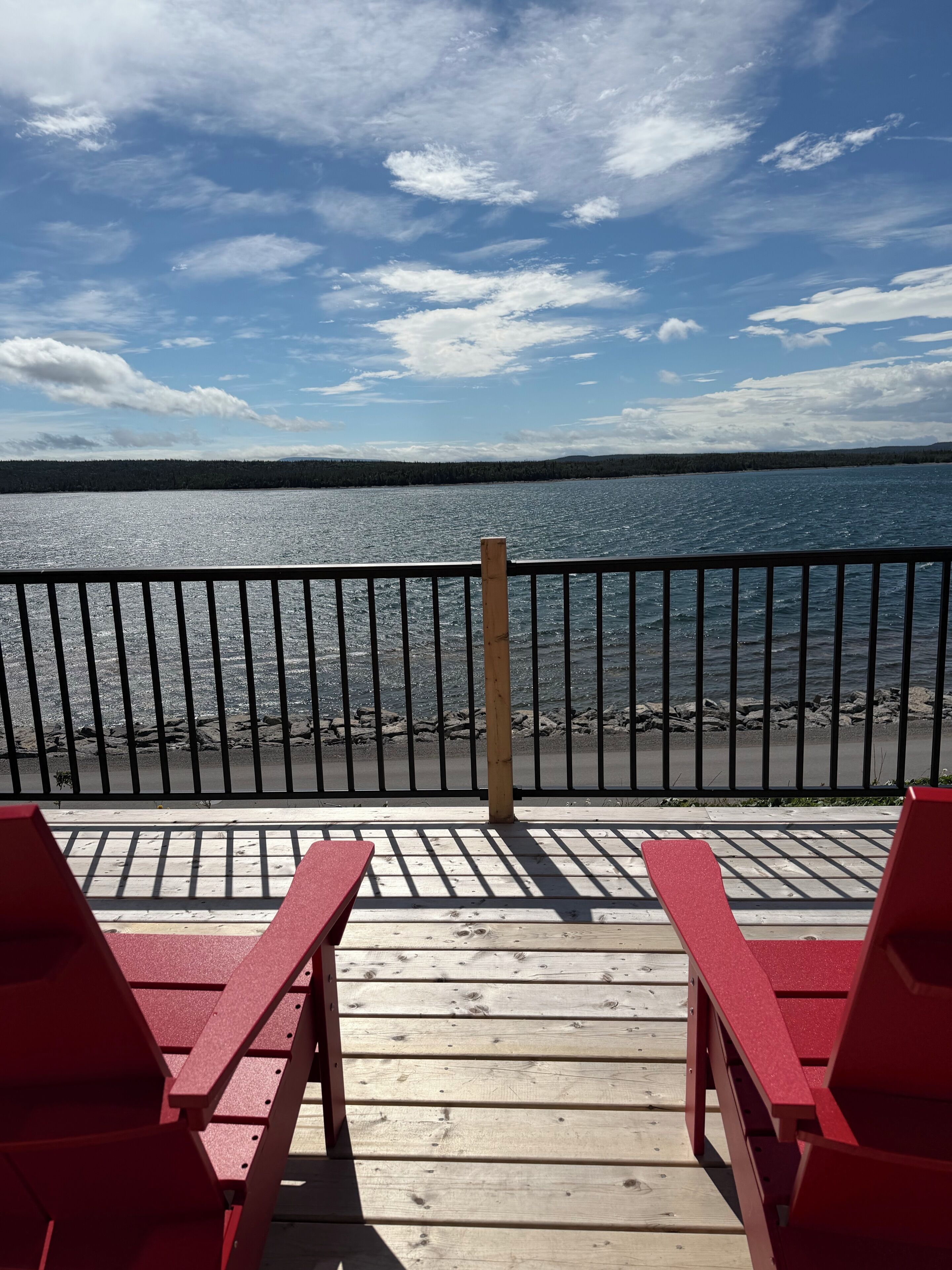 Une terrasse en bois avec deux chaises rouges donnant sur une étendue d’eau, une rambarde en métal et un ciel dégagé.