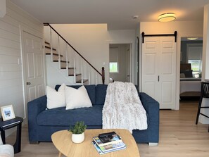A living room with a blue sofa, white pillows, a white fluffy throw, a coffee table with a plant and books, a staircase, and a door leading to a bedroom. - Saltwater Steeple- Bell Tower Suite (Port Saunders)