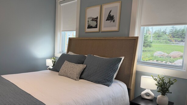 A bedroom with a bed, two framed pictures, a window with a view, and a bedside table with a plant. - Saltwater Steeple- Bell Tower Suite (Port Saunders)