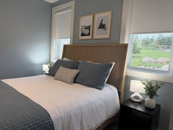 A bedroom with a bed, two framed pictures, a window with a view, and a bedside table with a plant.