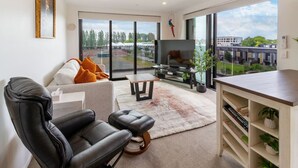 A modern living room with a sofa, a black leather chair, a TV, and a large window offering a view of buildings and trees. - Manchester Square Urban Stay – Easy To Everything (Christchurch)