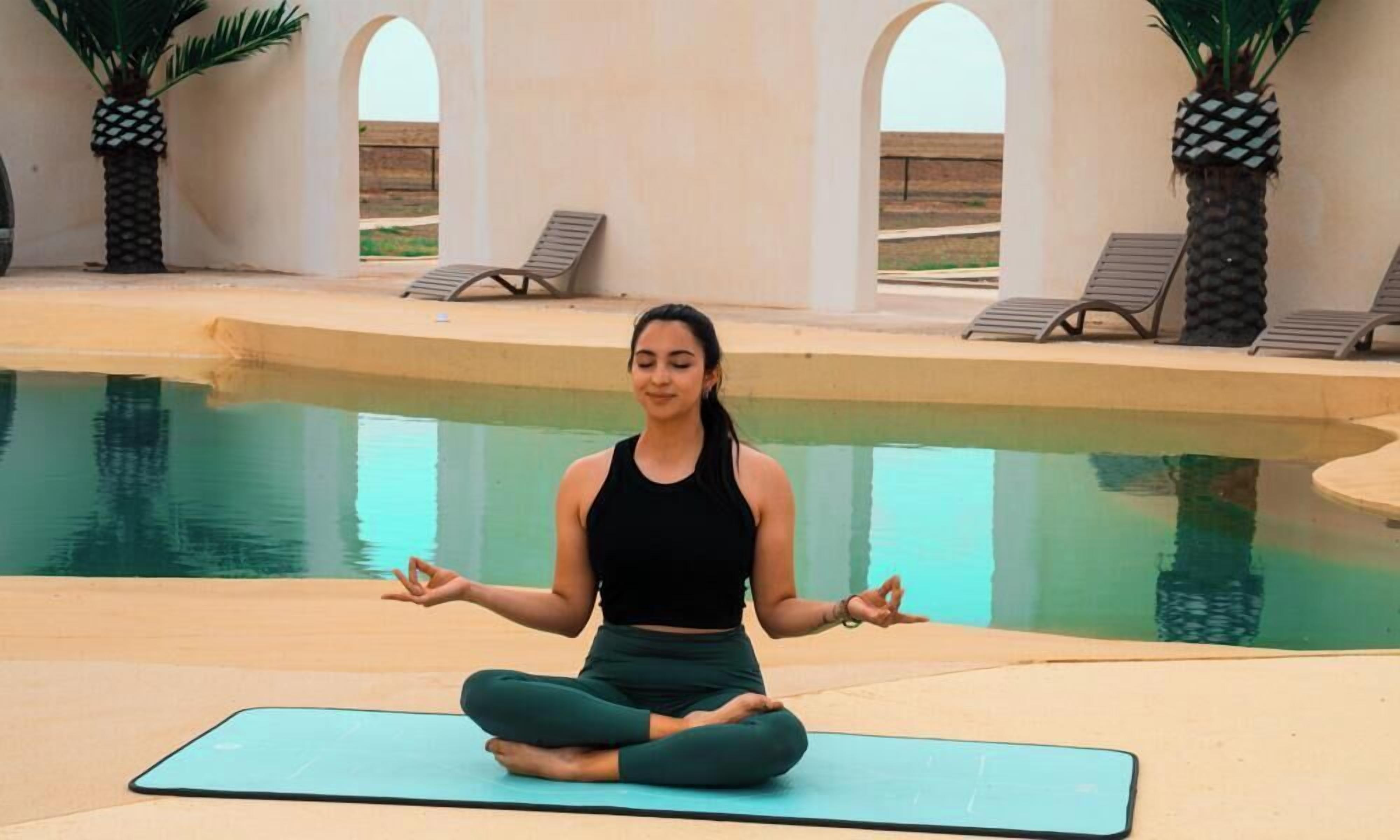 A person meditating on a mat by a pool with lounge chairs and palm trees in the background.