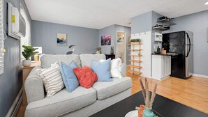 Ein modernes Wohnzimmer mit grauem Sofa, einer Küchenzeile mit schwarzem Kühlschrank und einem Esstisch mit Stühlen.