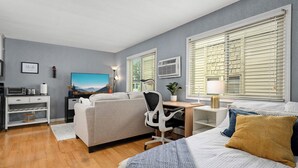 Ein modernes Hotelzimmer mit Bett, Schreibtisch, Stuhl, Sofa, Fernseher und einem Fenster mit Jalousien.
