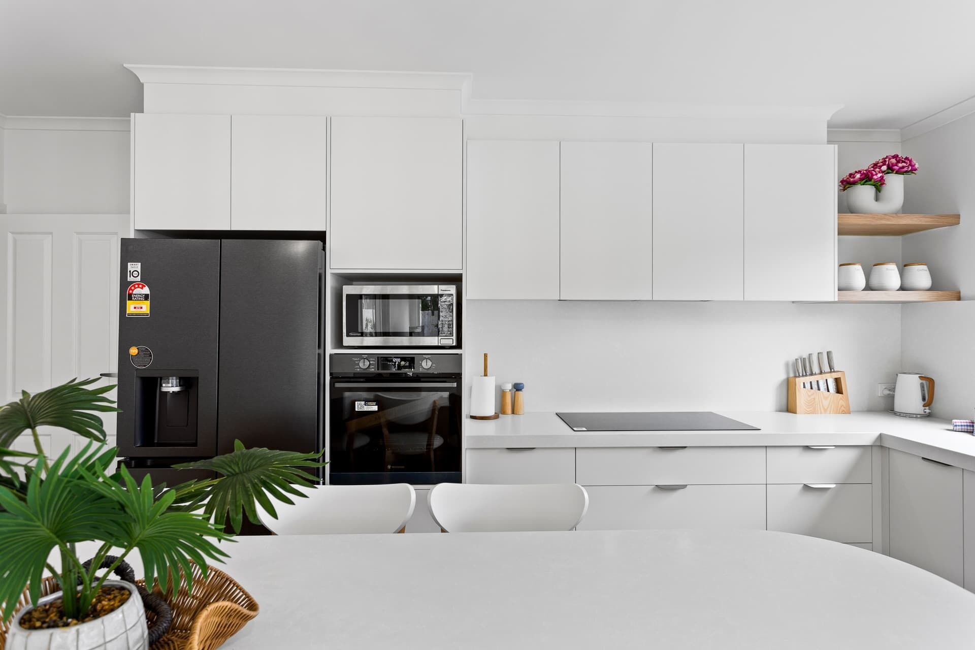 Eine moderne Küche mit weißen Schränken, einem schwarzen Kühlschrank, einer Mikrowelle und einem Backofen. Ein Esstisch mit Stühlen, ein Blumentopf und eine kleine Ablagefläche mit Dekorationen sind ebenfalls vorhanden.