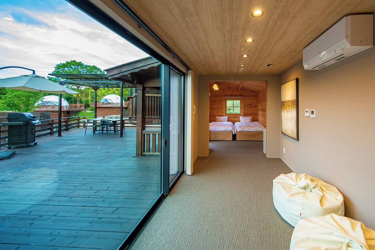 Een kamer met een houten plafond, een bed, een bankje en uitzicht op een terras met een barbecue en parasols.