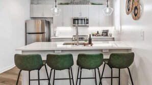 Eine moderne Küche mit weißen Schränken, einem Edelstahl-Kühlschrank, einer Mikrowelle und einem Spülbecken. Drei grüne Barhocker und Pendelleuchten befinden sich über der Kücheninsel.