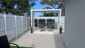 Un patio con un porche blanco, dos sillas colgantes y un pequeño jardín con grava y plantas.