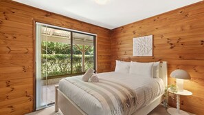 Une chambre avec un grand lit, des murs en bois, une porte-fenêtre coulissante donnant sur un jardin et une œuvre d’art murale blanche.