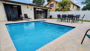 Ein rechteckiger Pool mit Fliesenrand, umgeben von einer Terrasse mit Stühlen und einem Tisch.
