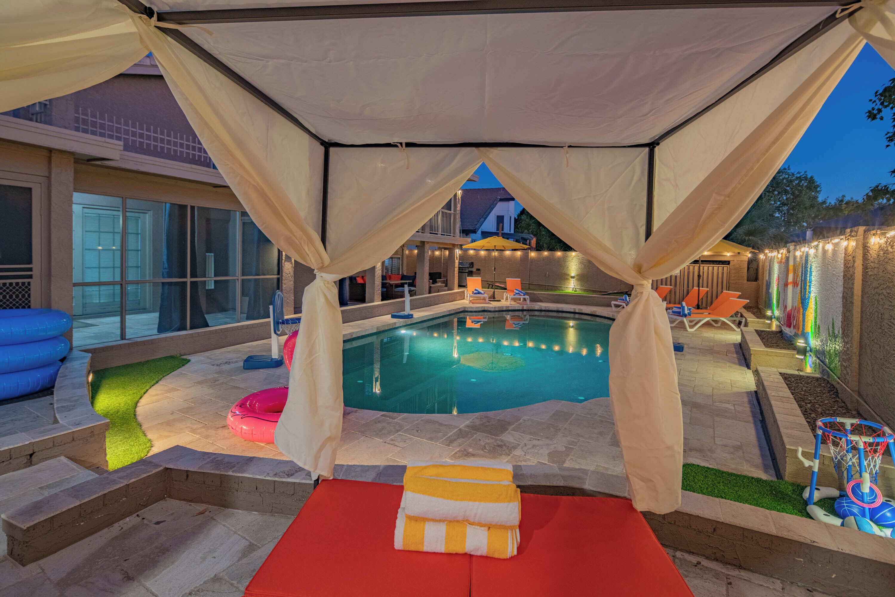 A swimming pool with a canopy, lounge chairs, and a small basketball hoop.