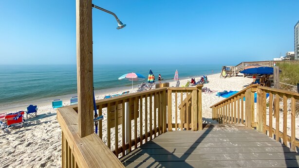 Eine Holzterrasse mit Metallgeländer, die auf einen Strand mit Sonnenschirmen und Liegestühlen blickt.