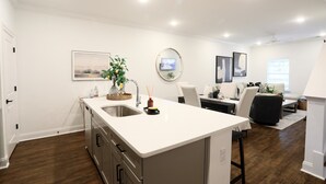 Une cuisine moderne avec un îlot central, un coin repas comprenant un canapé et une table basse, et un salon avec un divan et un téléviseur.