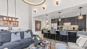Un salon moderne avec un canapé gris, une table basse ornée d’un vase et un coin repas avec des tabourets de bar.