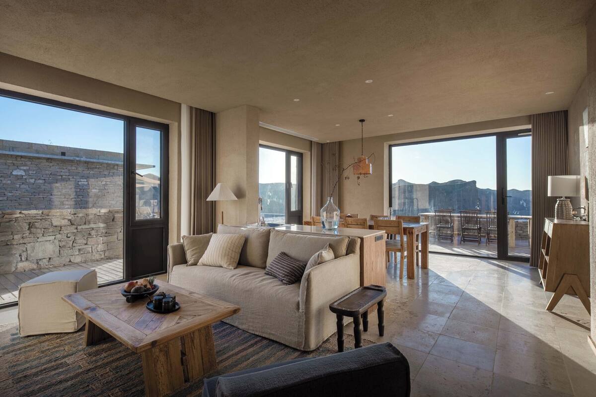 A spacious living area with a large sofa, coffee table, and dining set. The room features large windows offering a view of the mountains.