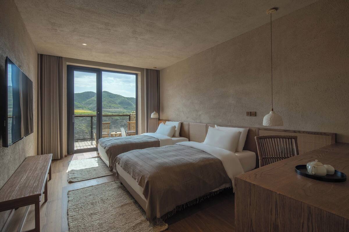 A modern hotel room with two beds, a desk, and a view of a mountainous landscape through large windows.