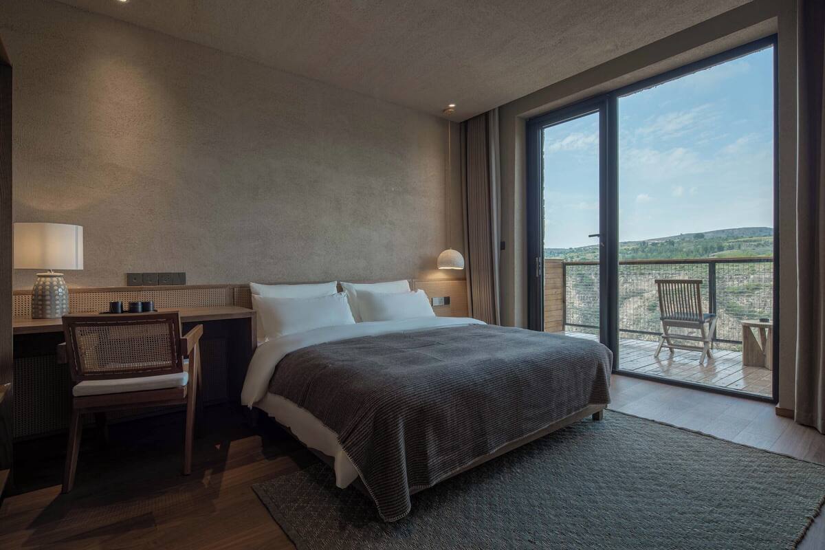 A modern hotel room with a large bed, a desk, and a chair. There is a balcony with a view of the countryside.