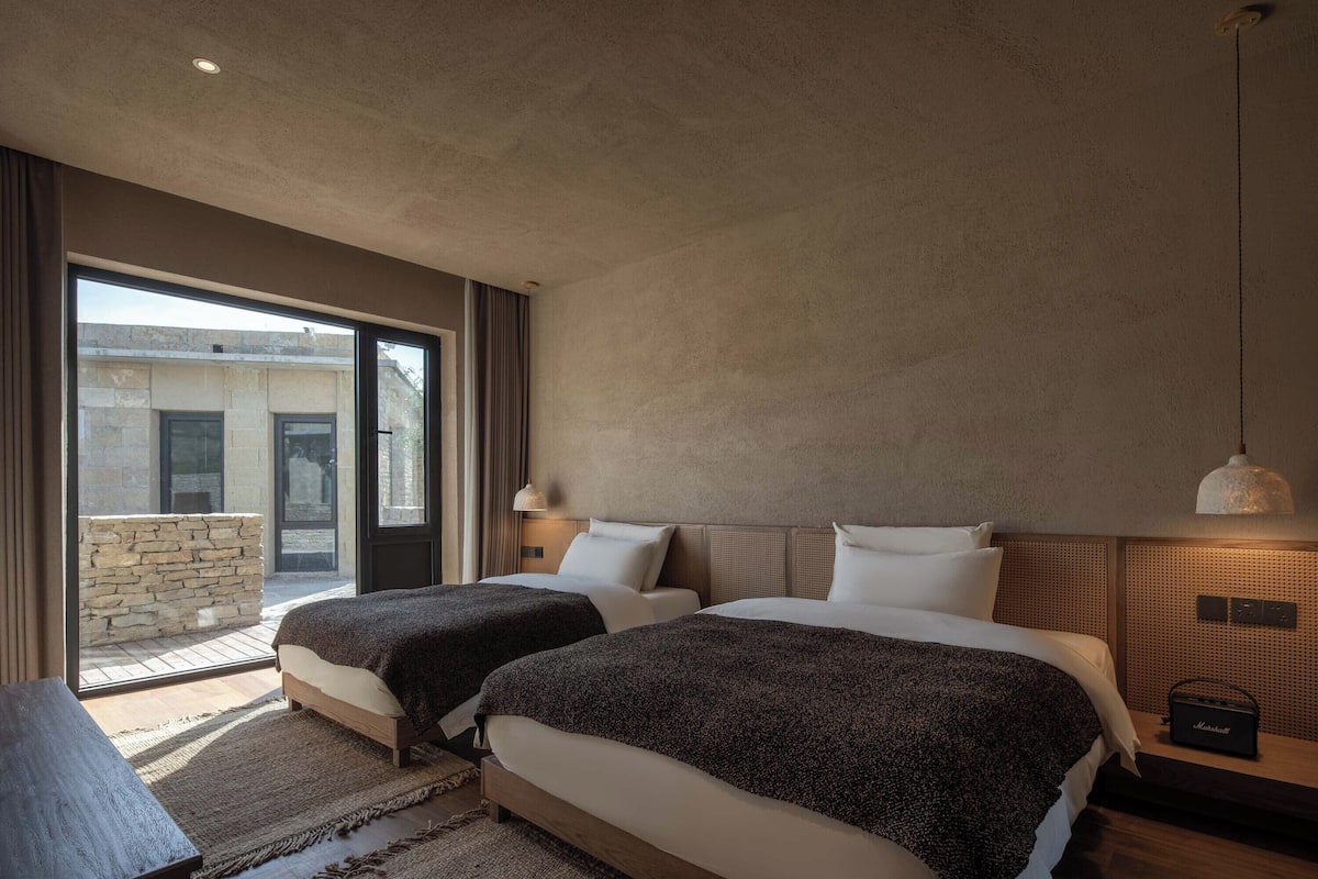A modern hotel room with two beds, a large window, and a stone wall outside.