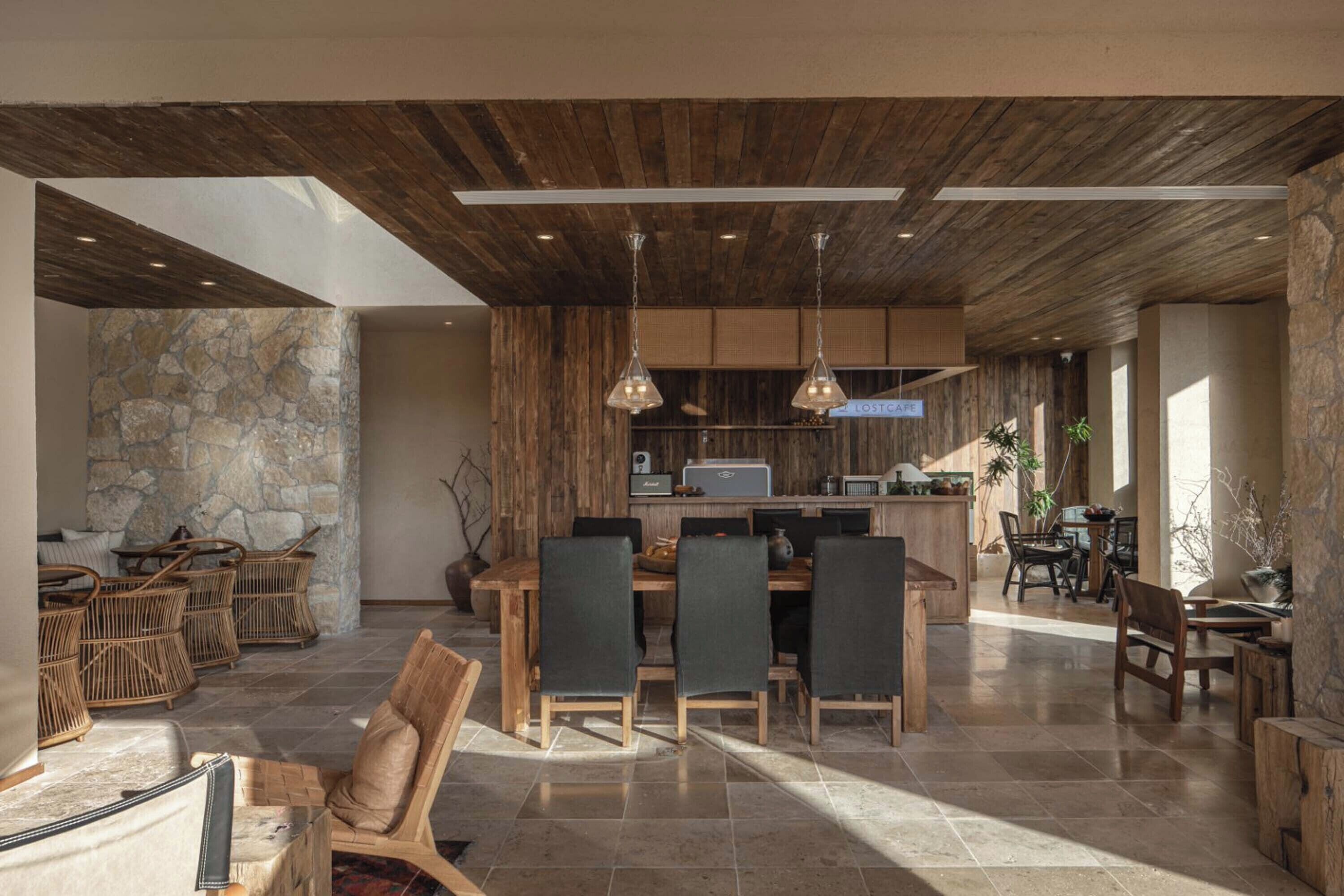 A spacious dining area with a stone wall and wooden ceiling, featuring a central table with chairs and a bar area with a computer and plants.