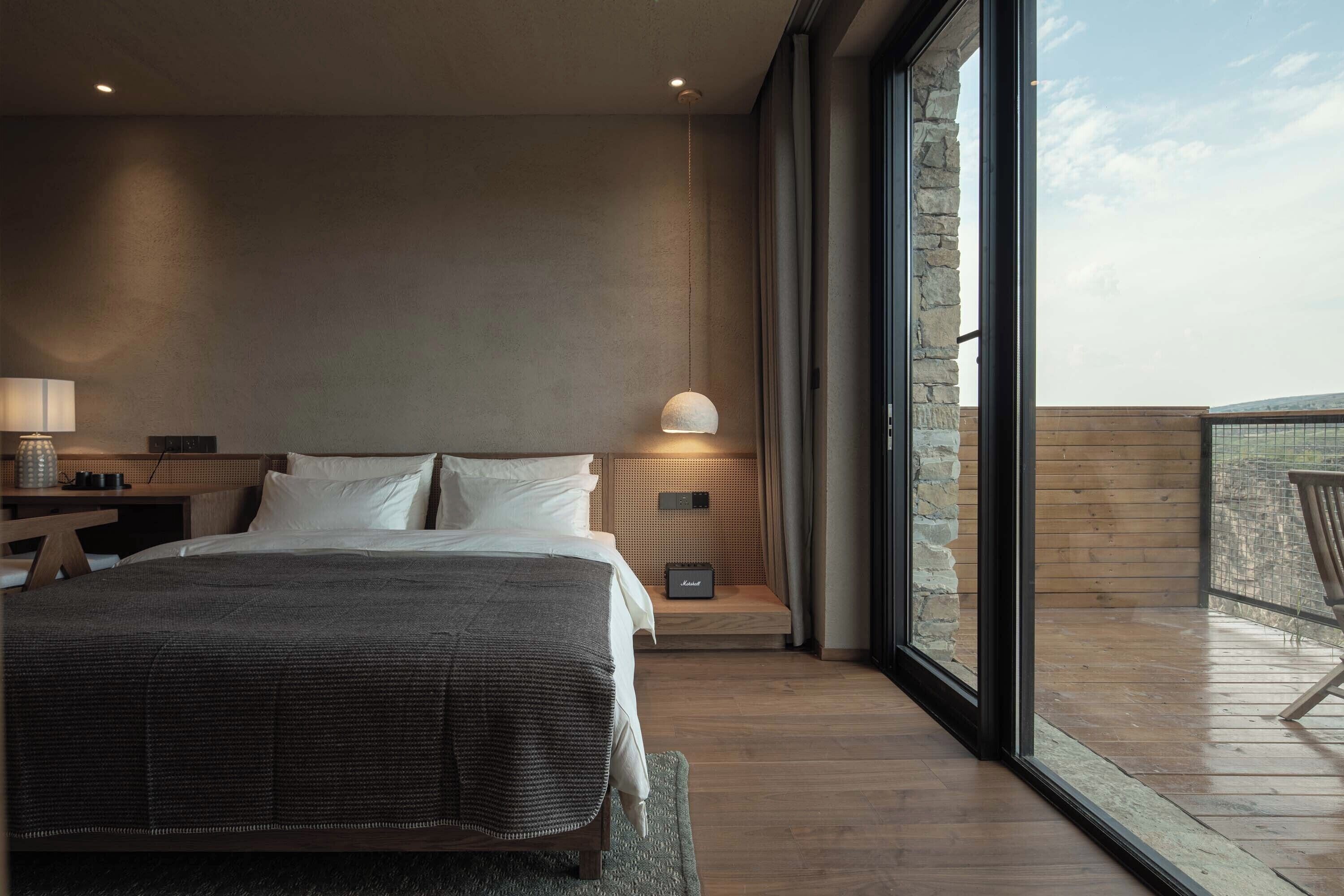 A modern bedroom with a large bed, a bedside table with a lamp, a small bedside table with a radio, and a view of the balcony.