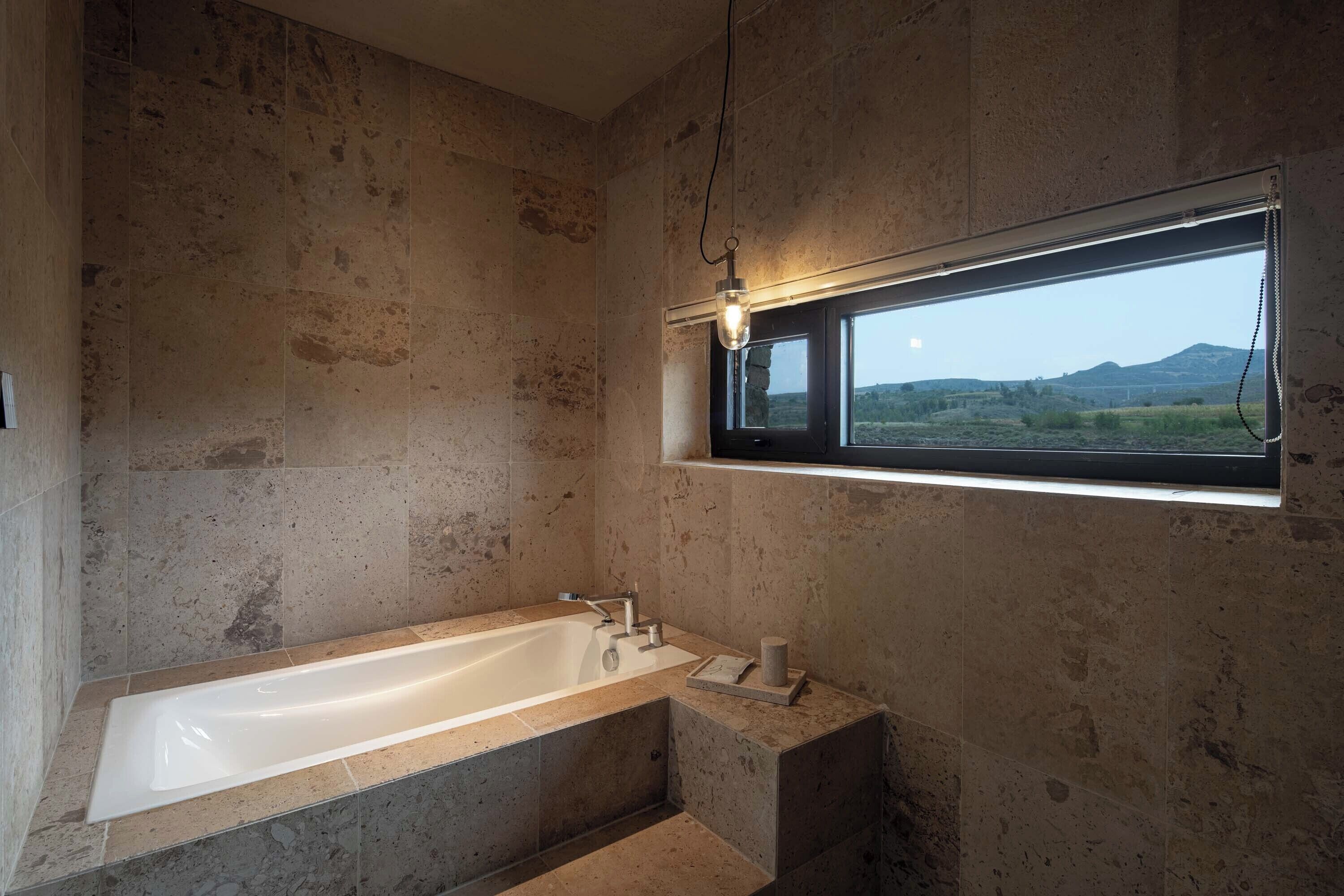 A modern bathroom with a large bathtub, a window with a view of the countryside, and a light fixture above the tub.