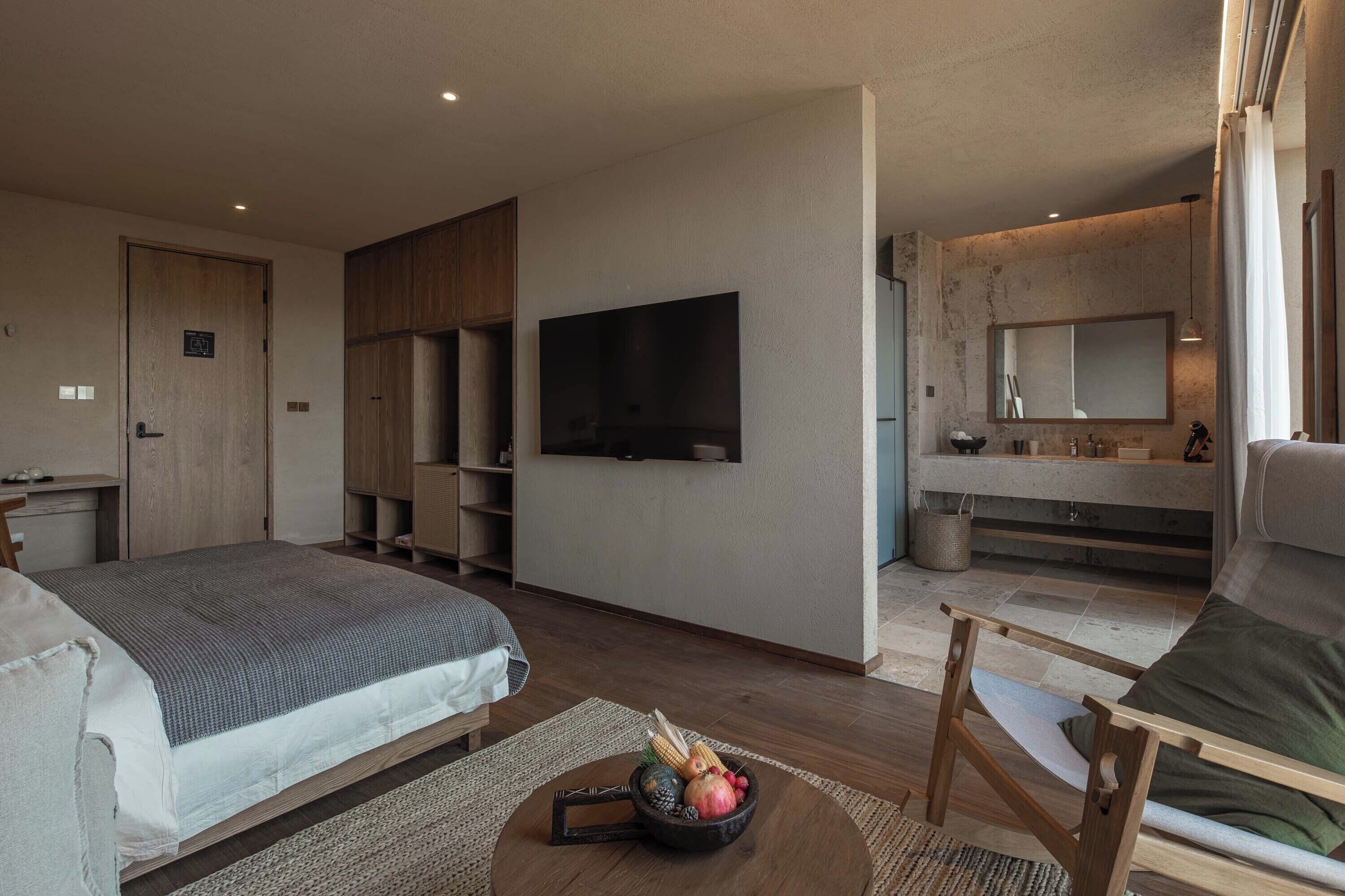 A modern hotel room with a bed, a TV, a small dining area, and a bathroom with a large mirror.