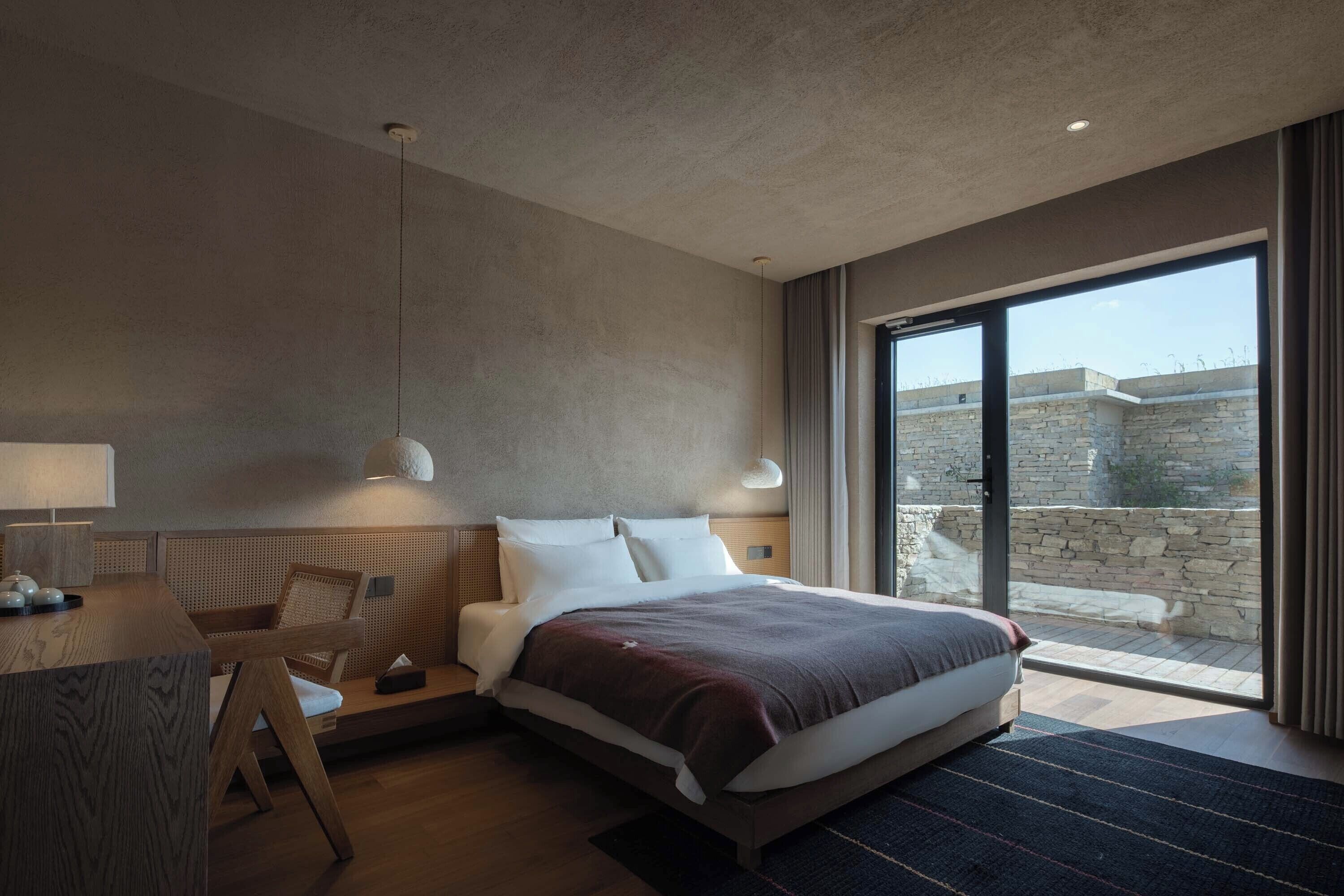 A modern hotel room with a large bed, a desk, and a view of a stone wall through a sliding glass door.
