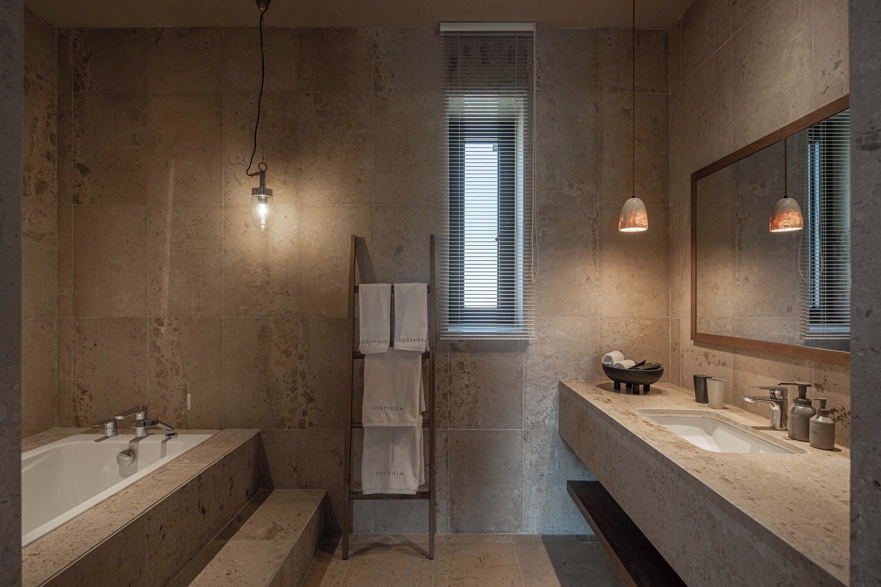 A modern bathroom with a large bathtub, a vanity with a sink, and a mirror. There are towels hanging on a rack and a window with blinds.
