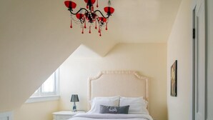 Une chambre avec un plafond en pente, un lit recouvert de draps blancs, une table de chevet avec une lampe et un lustre.