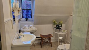 Une salle de bain équipée d’un lavabo, de toilettes et d’une douche. On y trouve un tabouret en osier et une fenêtre donnant sur des arbres.