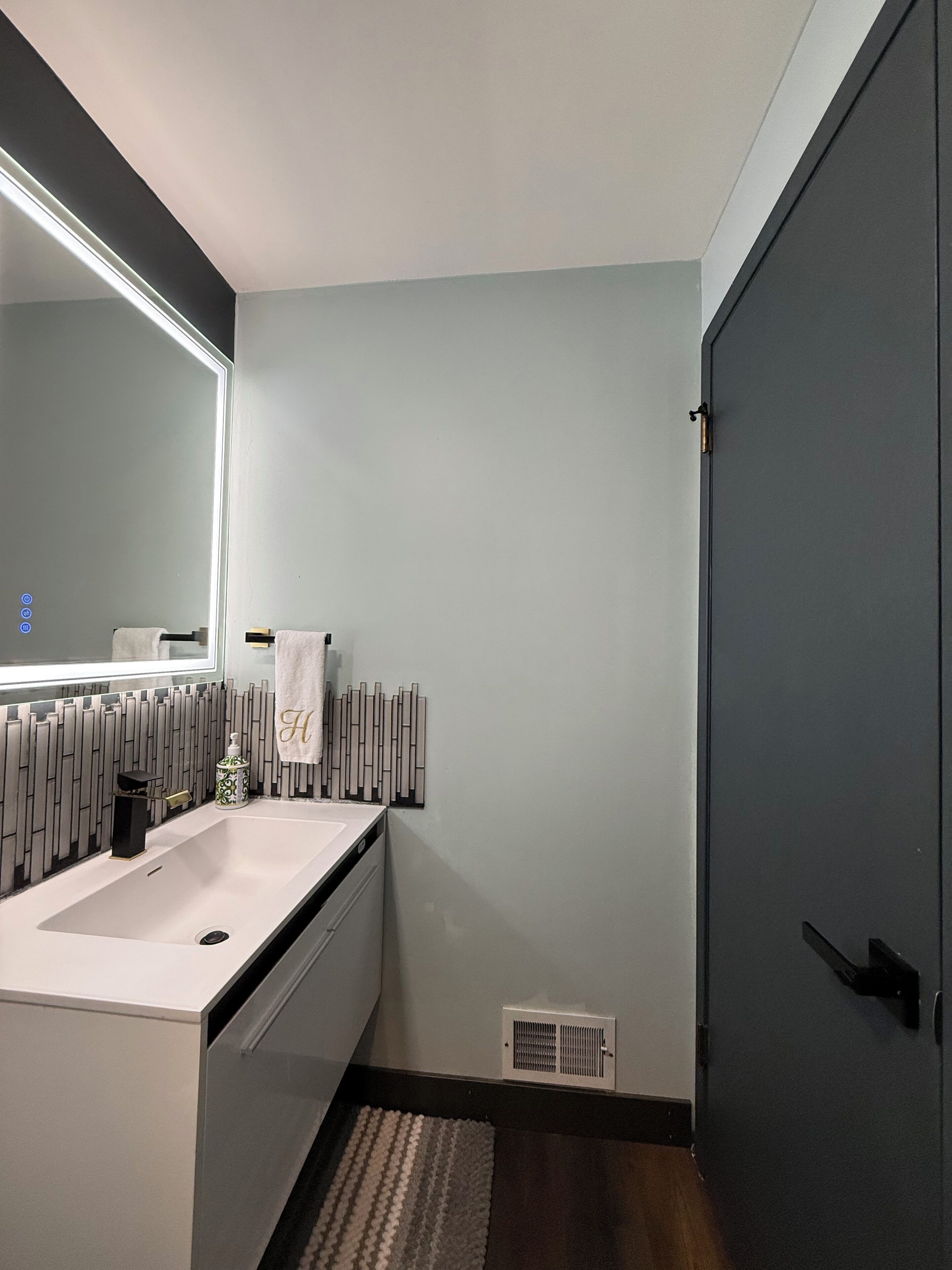 A modern bathroom with a large mirror, a white sink, a towel with a monogram, a radiator, and a door.