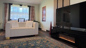 A living room with a white sofa, a large flat-screen TV, a fireplace, and a patterned rug. - Updated 3BR Home – Quiet, Comfortable Stay with Luxury Master & Spa Shower (Kennewick)