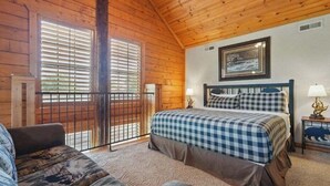 A cozy cabin bedroom with a bed, a chair, a window with blinds, and a picture on the wall. - Close to Shows, Dining & Golf • 2BR + Loft Cabin (Branson)