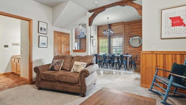 A cozy living room with a brown sofa, wooden beams, a dining area with a chandelier, and framed artwork on the walls. - Close to Shows, Dining & Golf • 2BR + Loft Cabin (Branson)