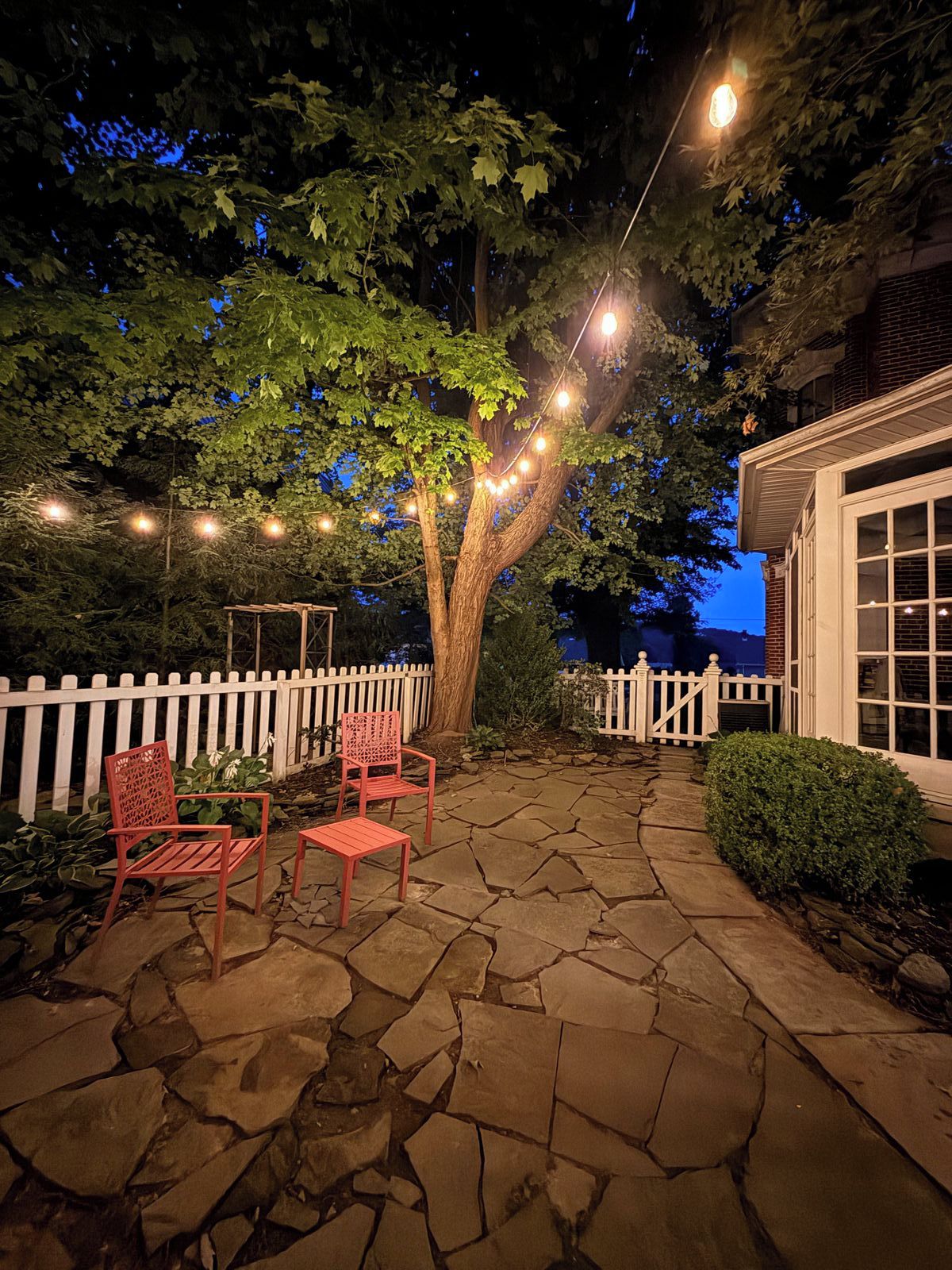 Un patio de piedra con dos sillas y una mesita pequeña, rodeado por una cerca blanca de piquetes y un árbol con luces colgantes.
