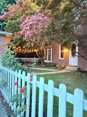 Een bakstenen huis met een witte schutting, een boom met roze bladeren en lichtjes.