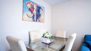 A dining area with a glass-top table, four chairs, a centerpiece, and a colorful elephant painting on the wall.