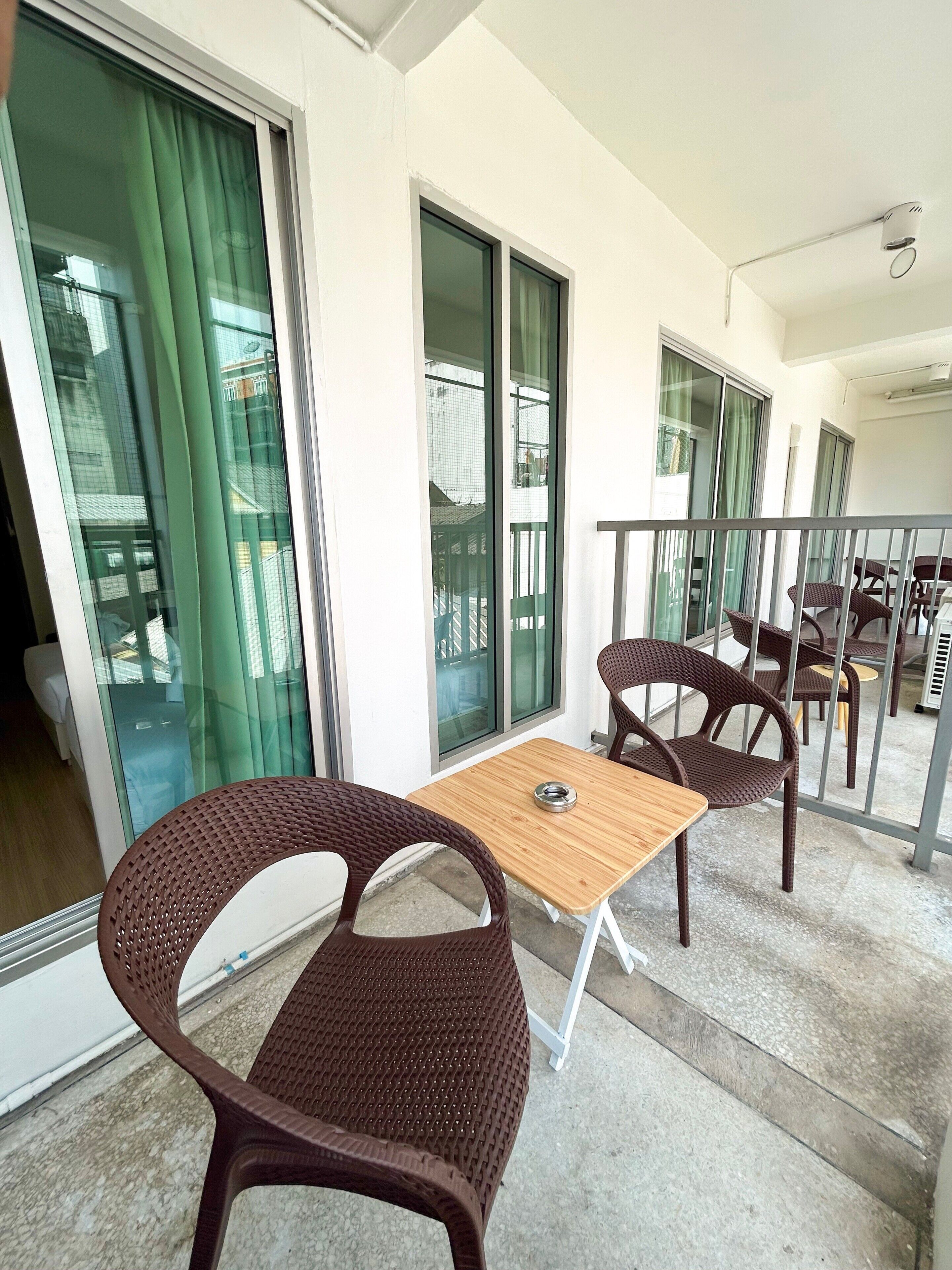 Un balcón con sillas de mimbre y una mesa, ventanas amplias y vista al interior del edificio.