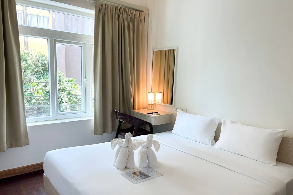 Une chambre d’hôtel avec un lit, un bureau, une chaise, une lampe et une fenêtre avec des rideaux.