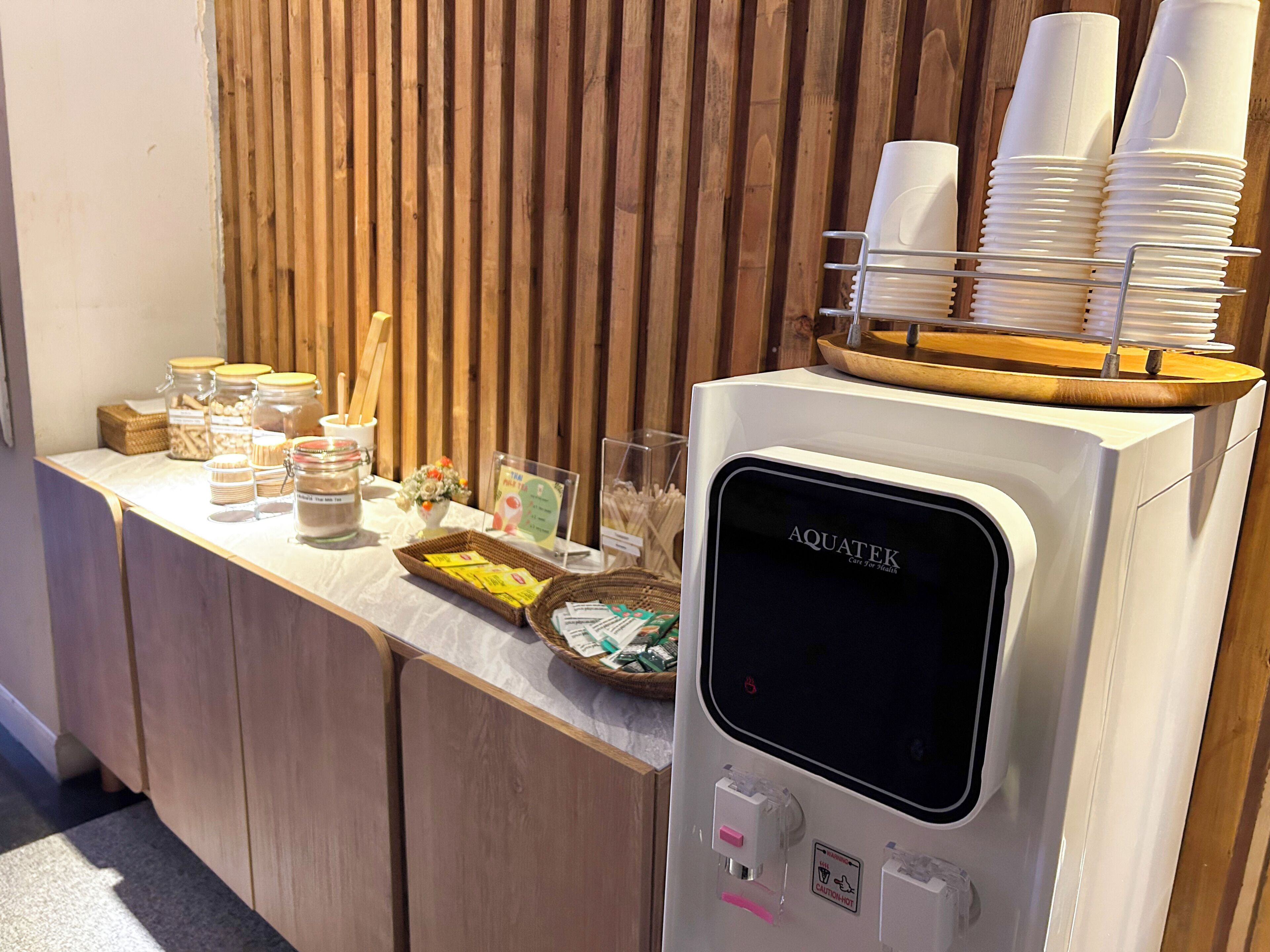 A self-service beverage station with a coffee machine, a tray of disposable cups, and various food items on a counter.