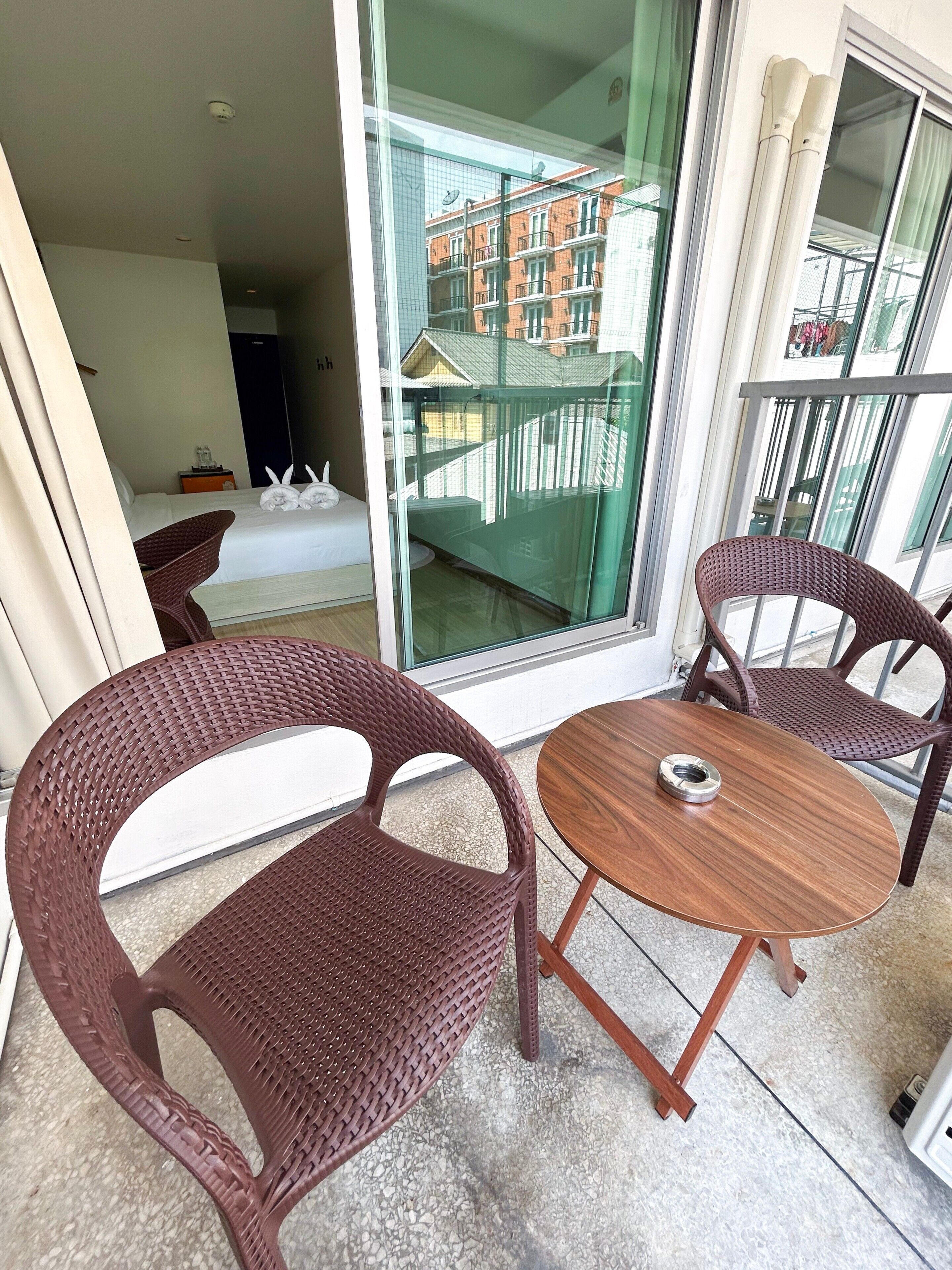 A balcony with wicker chairs and a small wooden table, a bed with white linens, and a view of a building with balconies.