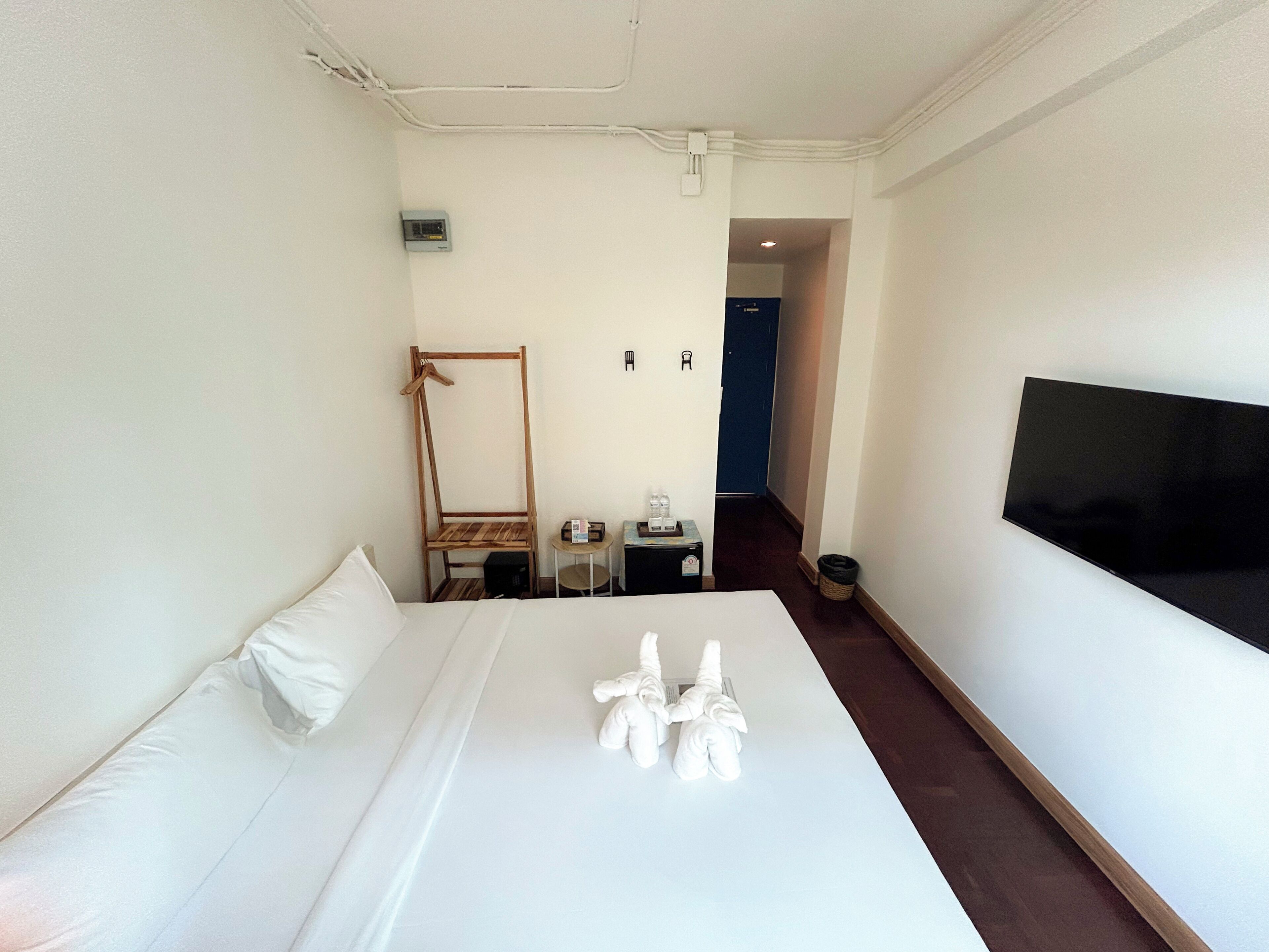 A hotel room with a bed, a wooden chair, a small table with a bottle, a trash bin, and a TV on the wall.