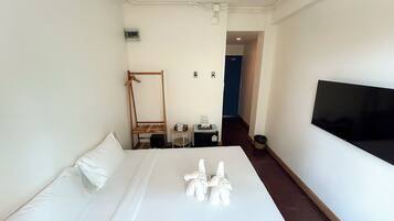 A hotel room with a bed, a wooden chair, a small table with a bottle, a trash bin, and a TV on the wall.