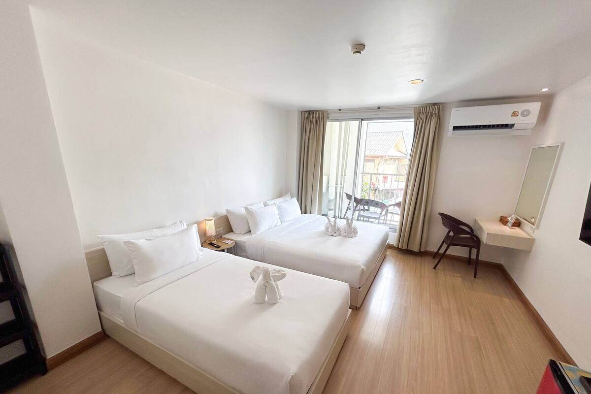 Une chambre d’hôtel avec deux lits, un bureau, une chaise, un appareil de climatisation et une fenêtre avec des rideaux.