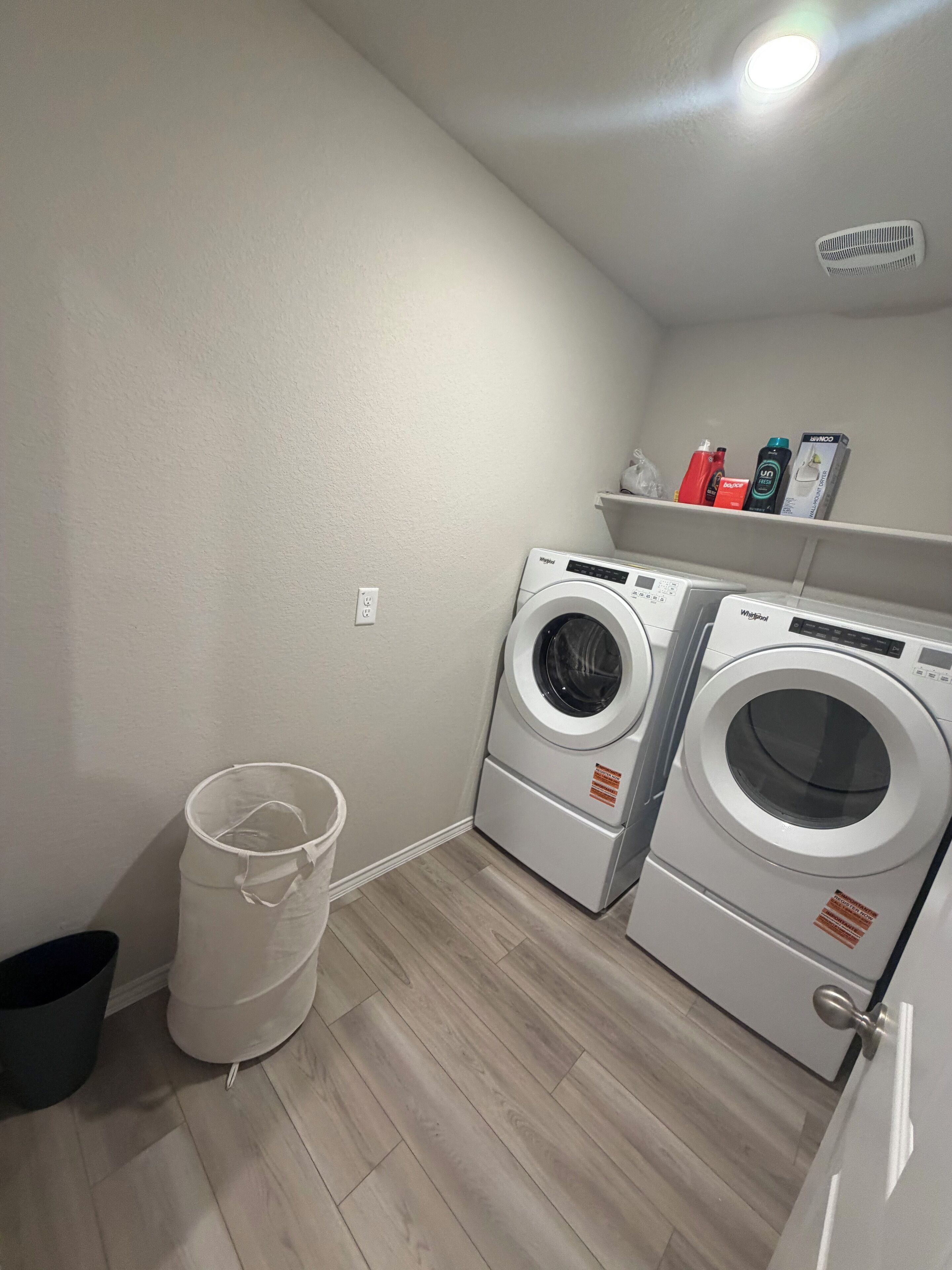 A laundry room with two front-loading washing machines, a white trash bin, and a shelf with cleaning supplies.