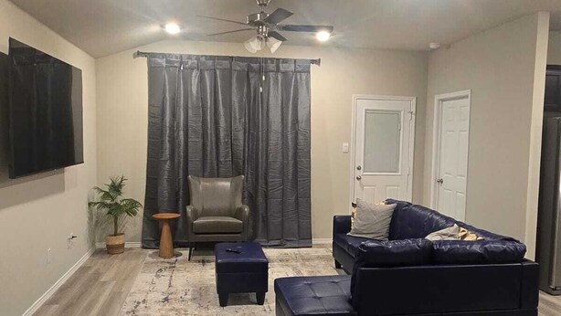 A living room with a dark blue sofa, a small ottoman, a chair, a ceiling fan, and a large window with curtains. - Contemporary 3BR Home Near Base Fast Wi-Fi, Smart TV, Coffee Station (Killeen)