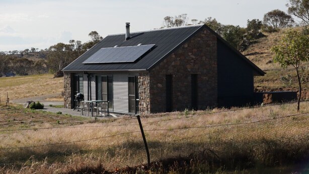 A stone building with a sloped roof and solar panels, surrounded by dry grass and sparse trees. - Skippy`s Cottage at Touchdown Cottages (Jindabyne)