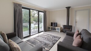 A living room with a sofa, a TV, a fireplace, and a sliding glass door leading to a patio. - Skippy`s Cottage at Touchdown Cottages (Jindabyne)
